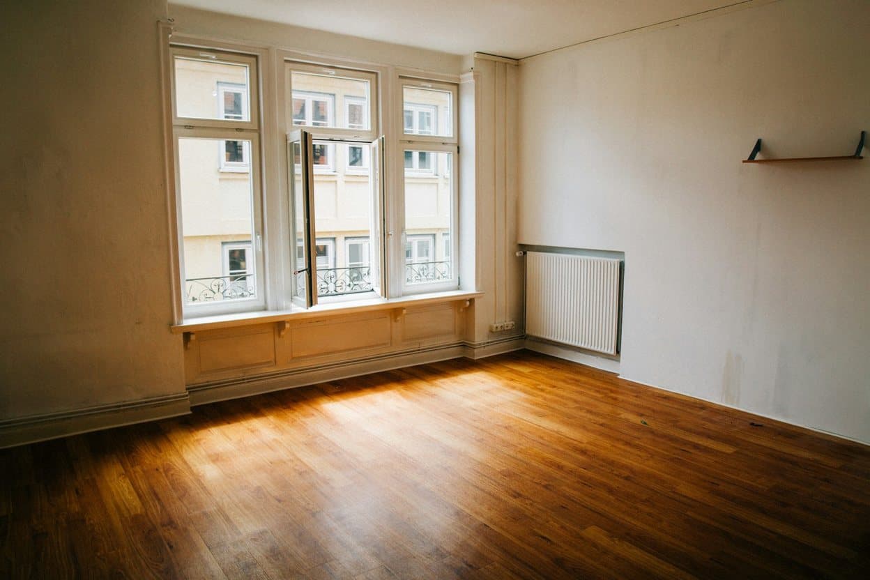 An empty apartment living room with tall windows, a radiator and warm oak floorboards